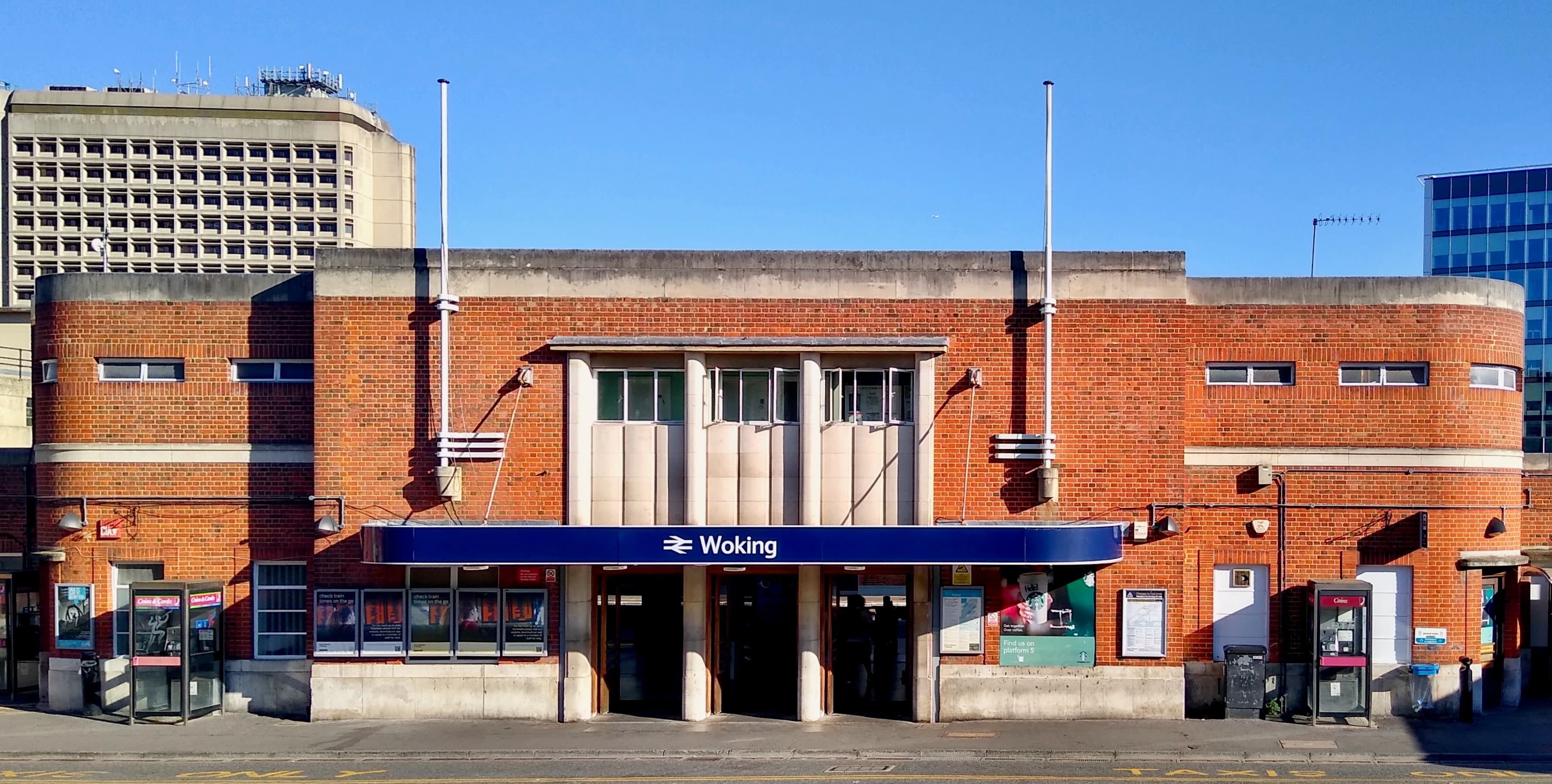 Red-brick art deco frontage of Woking railway station under a clear blue sky, with entrance canopy and phone boxes
