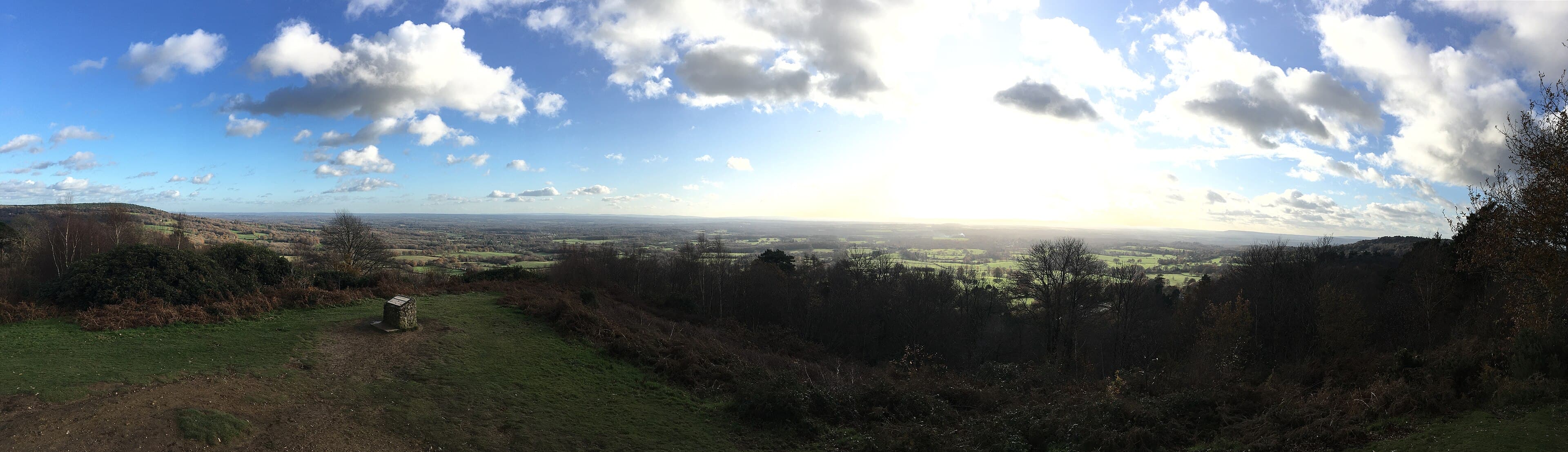 Panoramic view from a Surrey Hills heathland summit across wooded slopes and sunlit farmland under a bright, cloudy sky