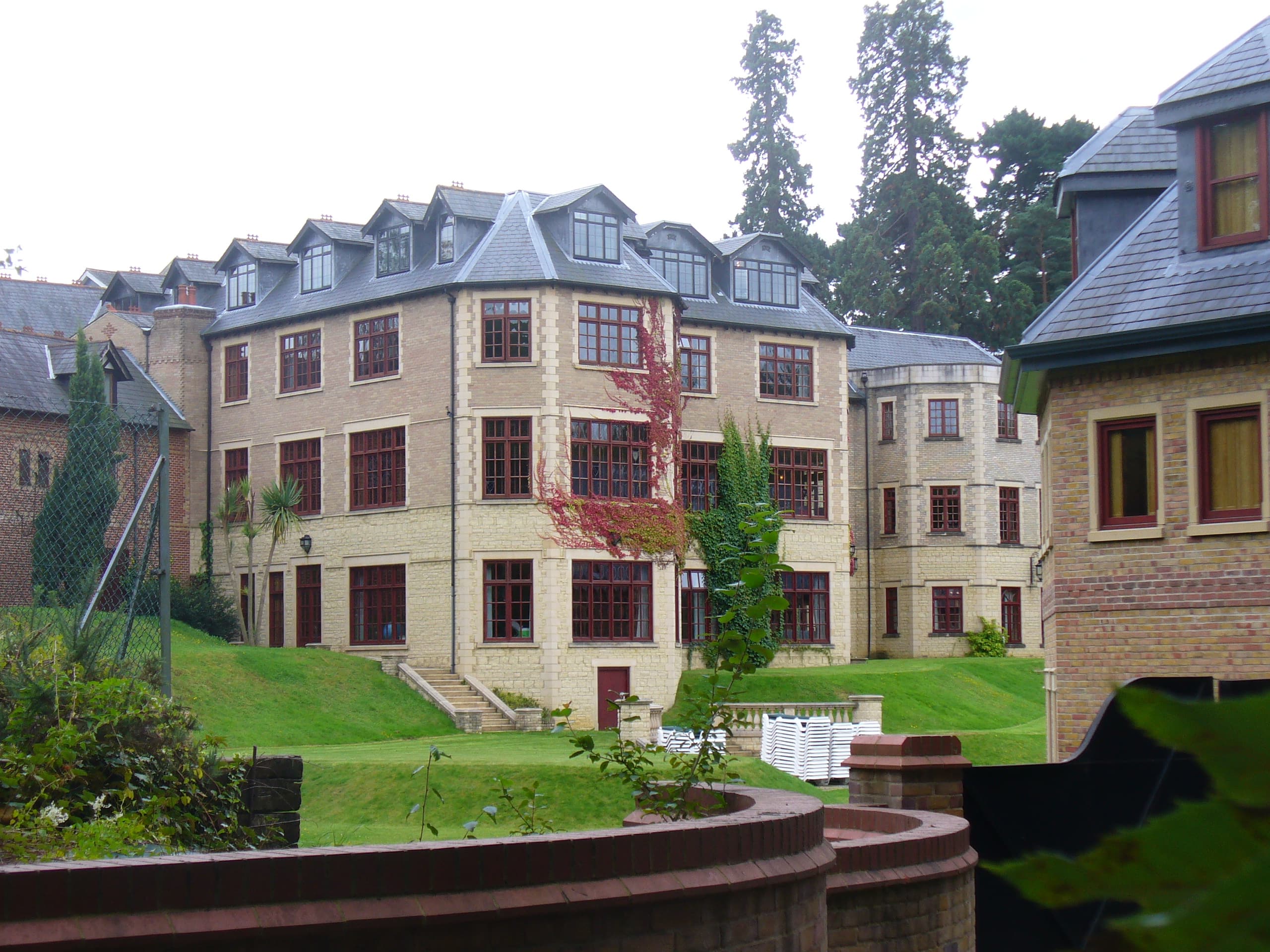 Large Victorian country house with slate dormer roof, stone and brick walls, red-framed windows and terraced lawns beside tall conifers