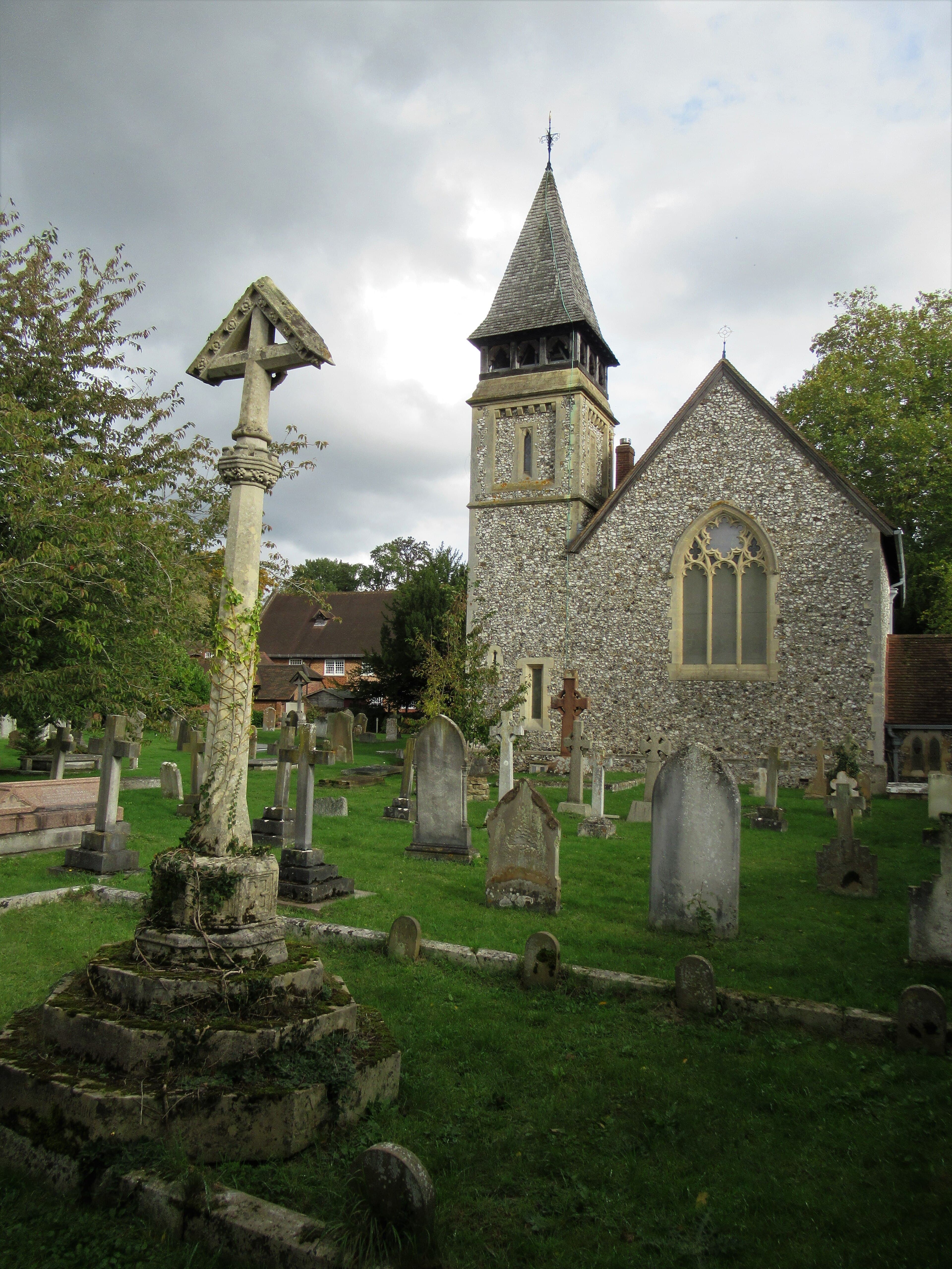 Flint-walled village church with shingled spire and Gothic window, seen across a graveyard of crosses and weathered headstones