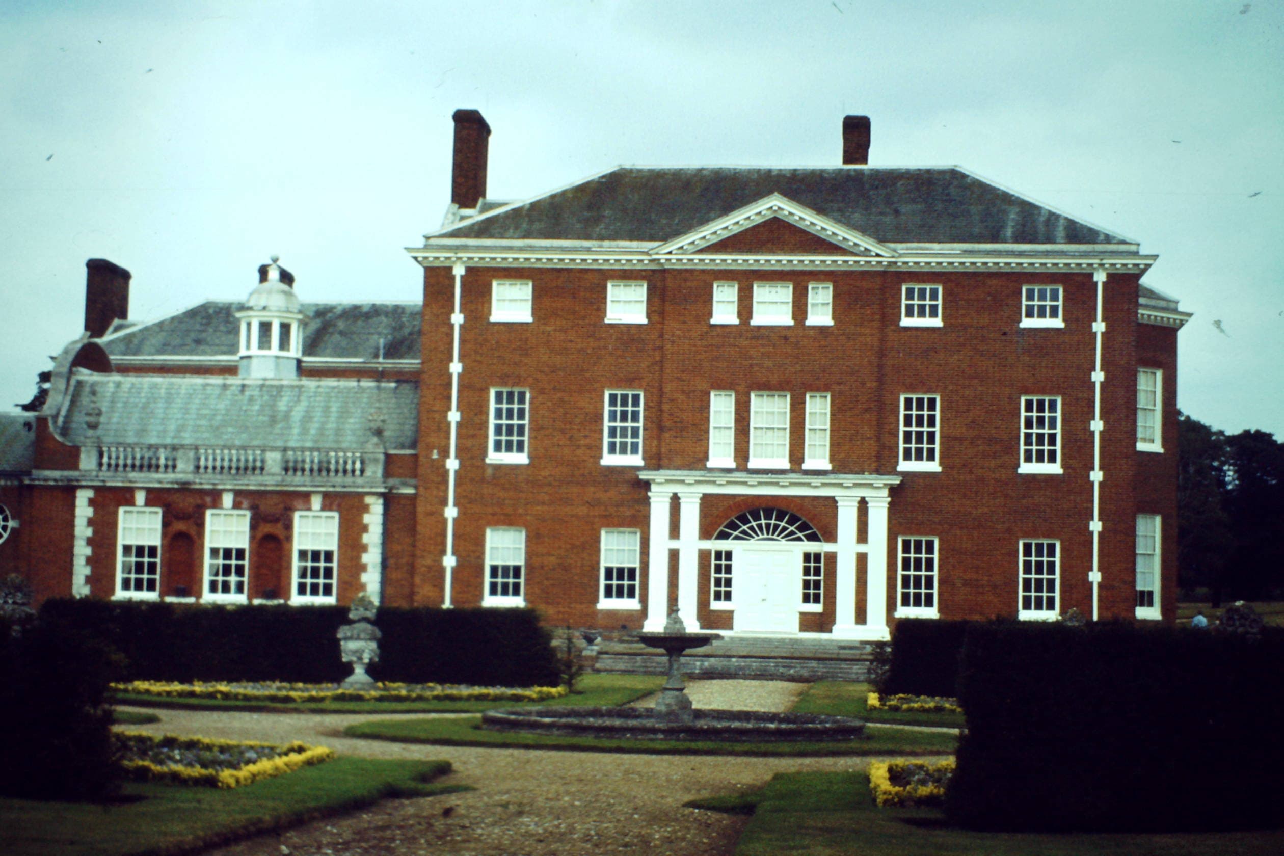 Red-brick Georgian country house with white portico, sash windows and formal gardens with a fountain under an overcast sky