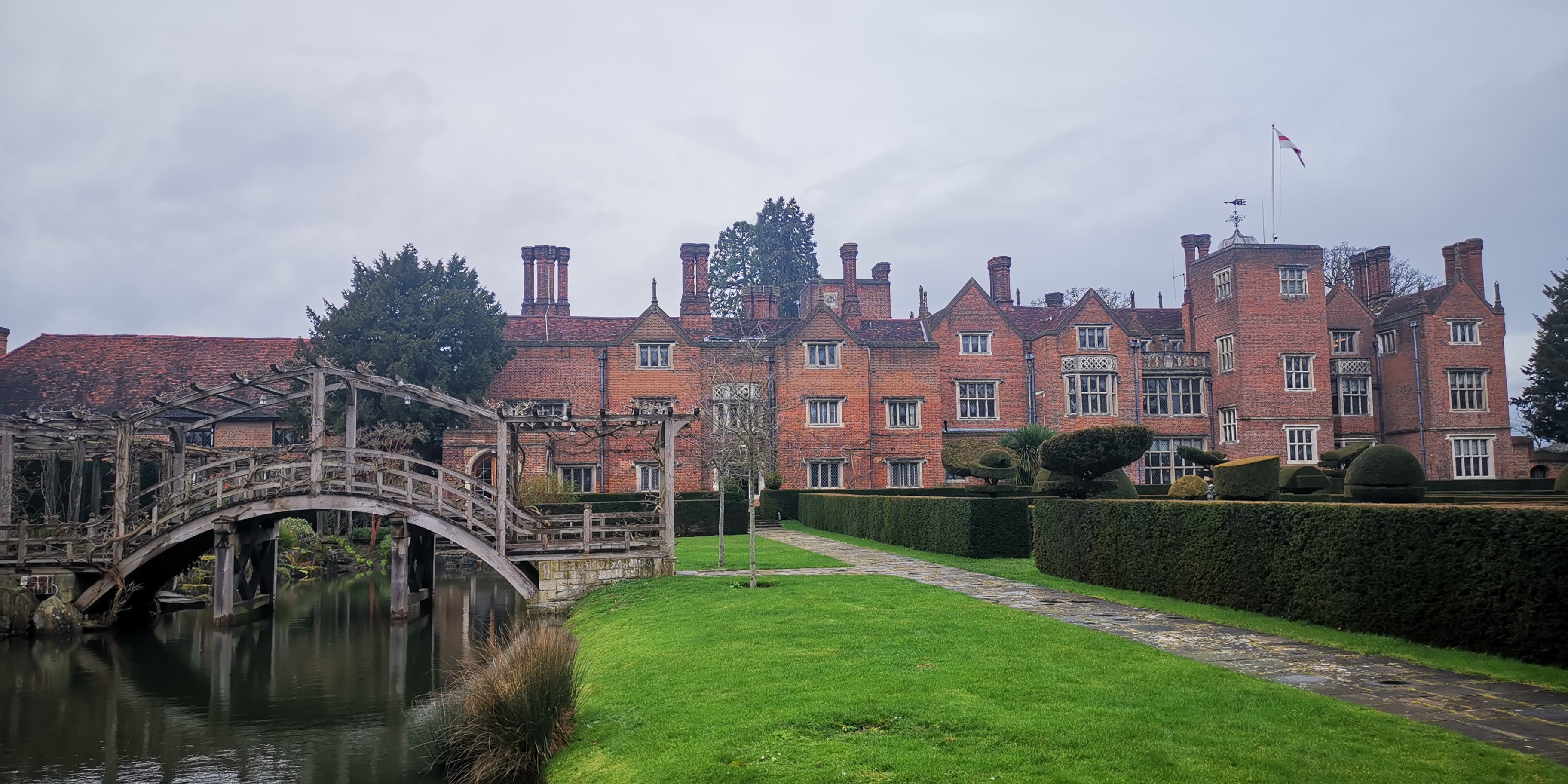 Tudor red-brick country house with tall chimneys, clipped topiary, lawn and a wooden arched bridge over a pond