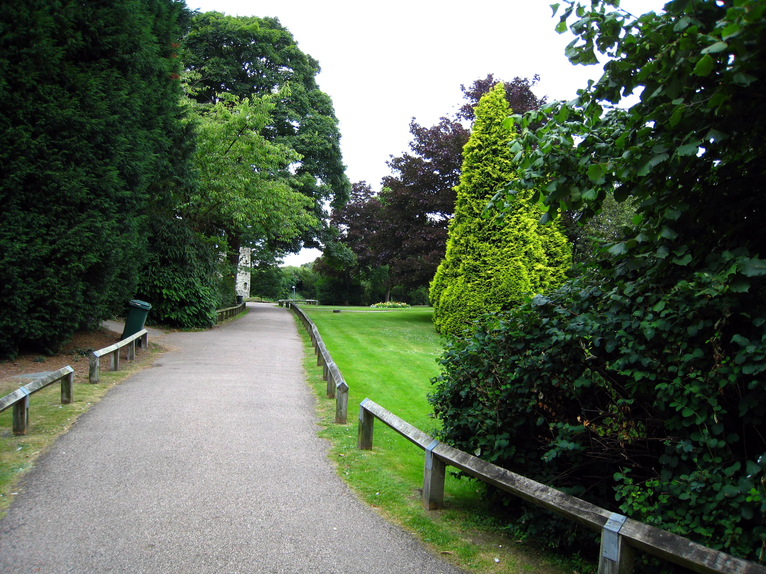 Tarmac path beside a mown lawn and wooden rails, with mature trees and a stone tower beyond