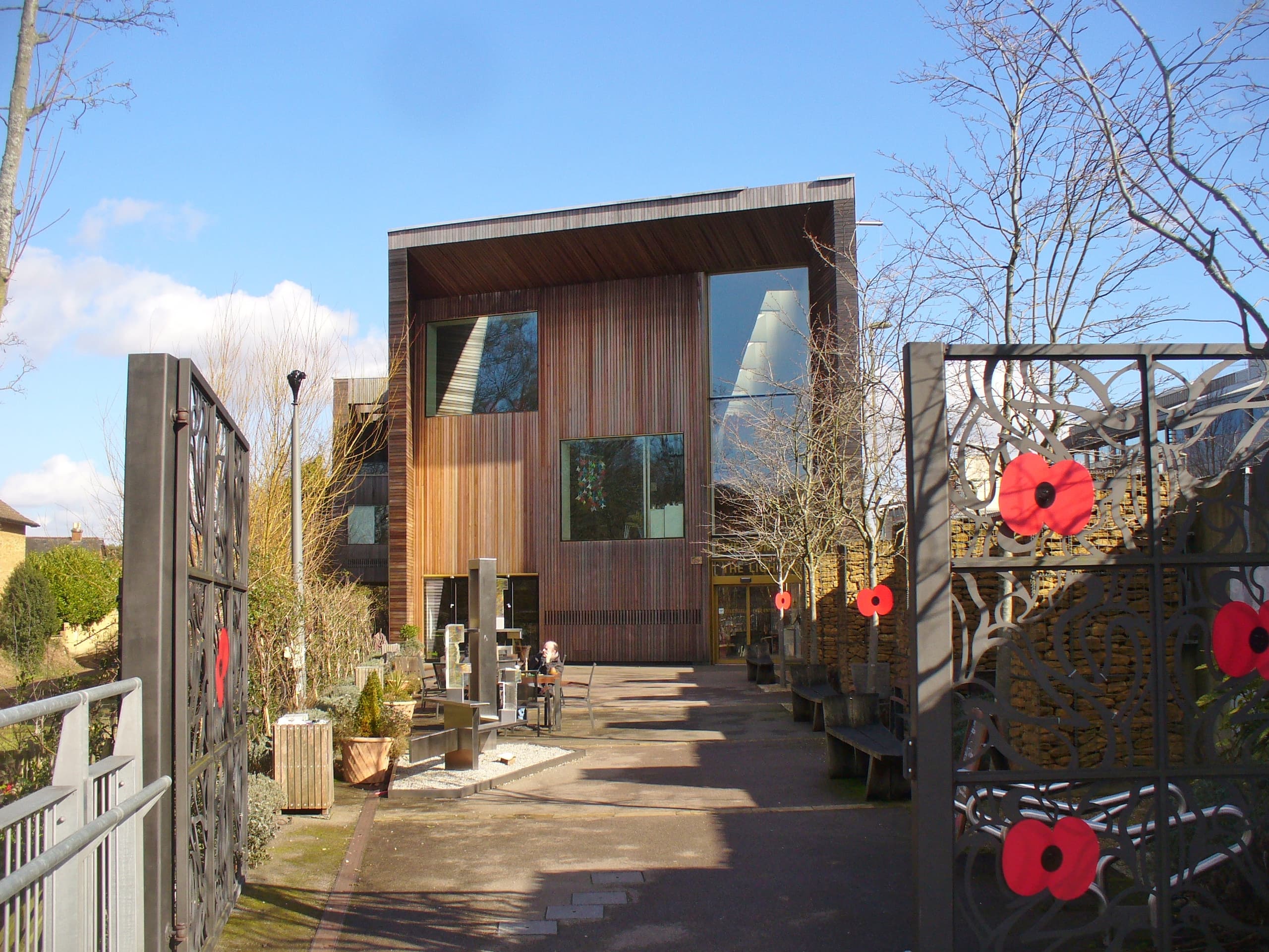 Timber-clad Lightbox gallery in Woking seen through ornate metal gates decorated with red poppies, under blue sky