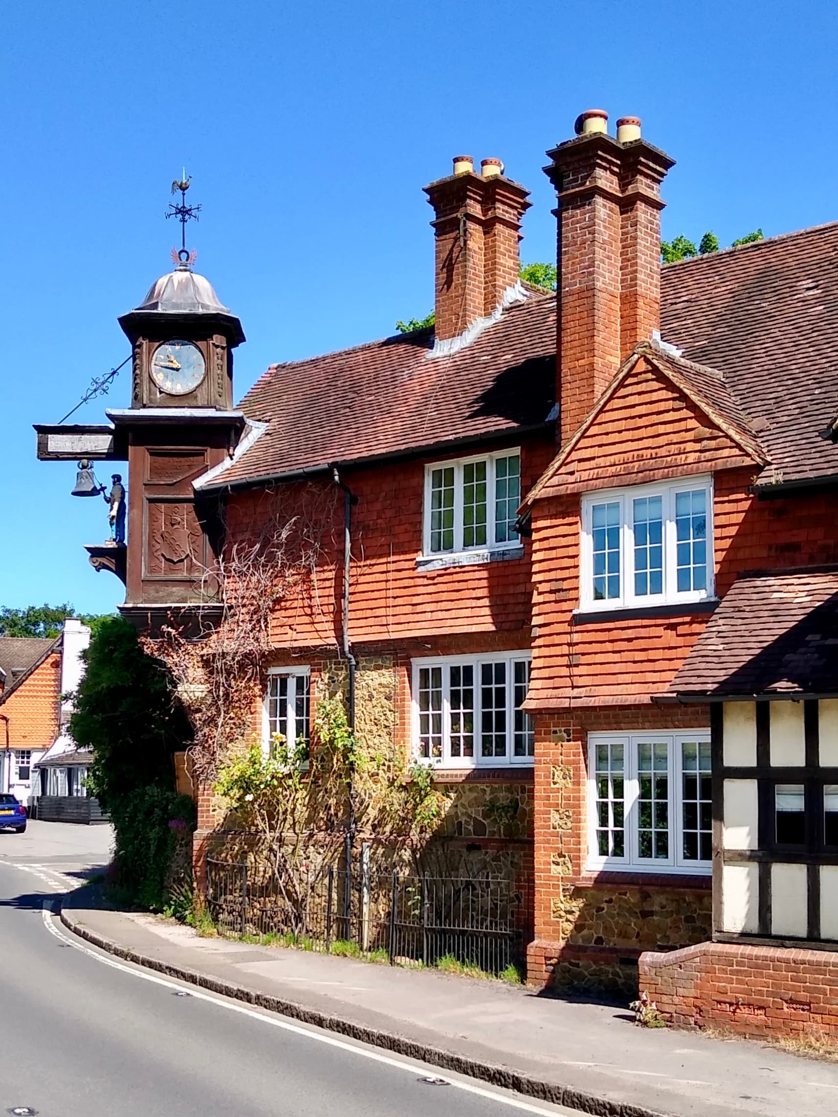Abinger Hammer's clock tower with bell and blacksmith figure projecting over the road beside red-tiled cottages under blue sky