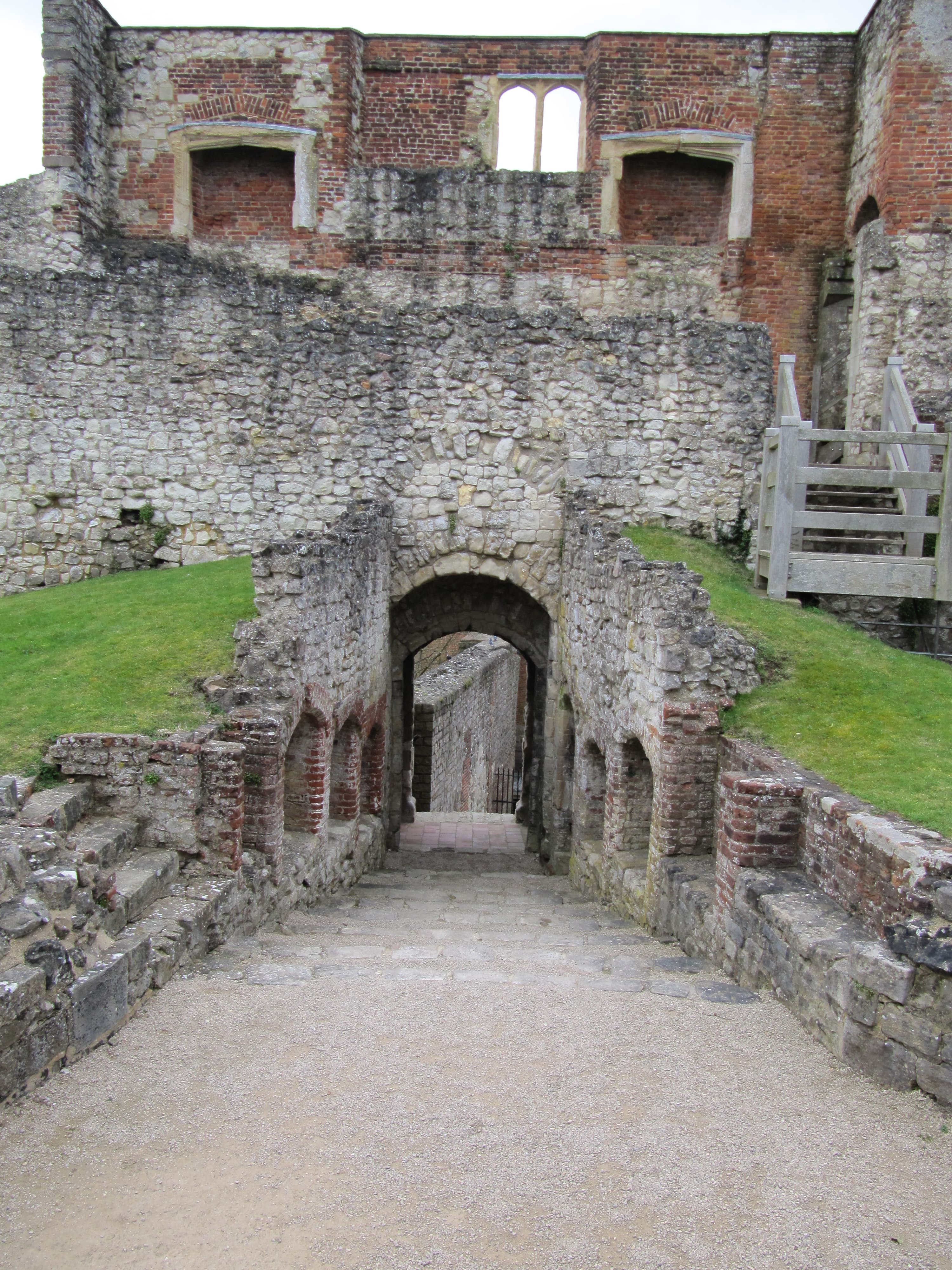 Flint and red brick ruined castle walls with an arched stone passage descending from a gravel path beside grassy banks