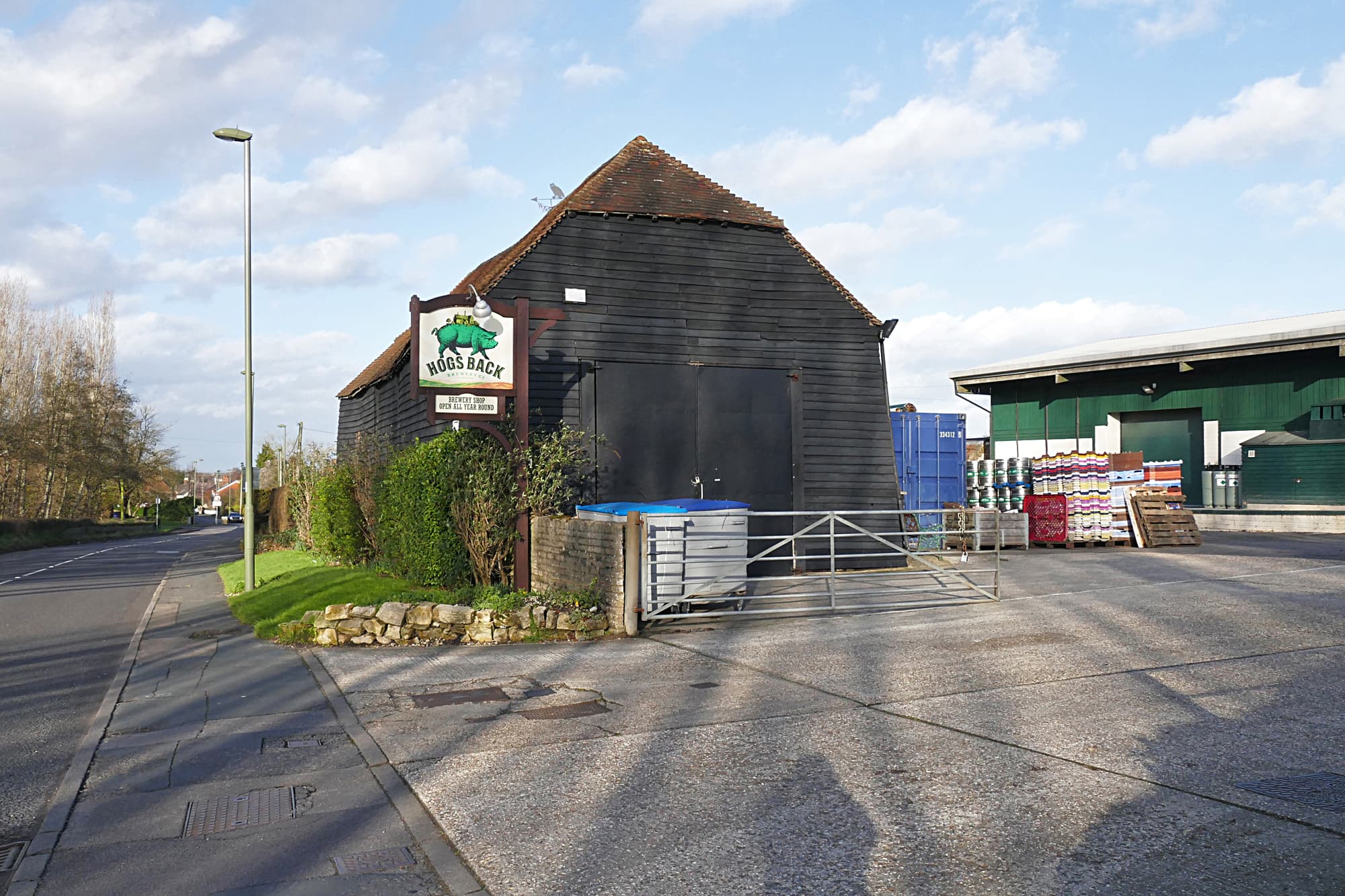 Black weatherboarded barn with Hogs Back Brewery sign, kegs and pallets stacked beside a green warehouse in Tongham