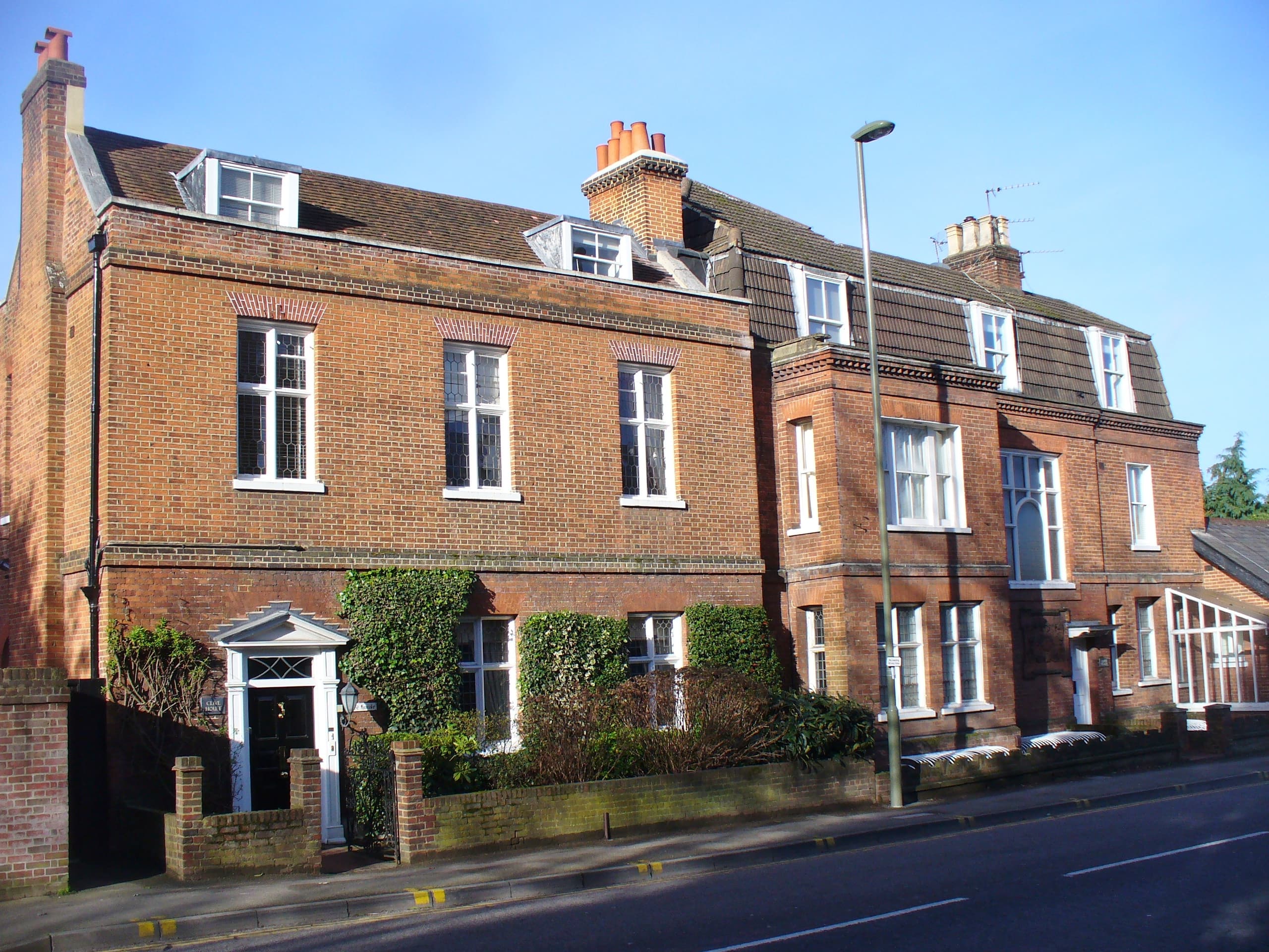 Red brick Georgian and Victorian townhouses with dormer windows and tall chimneys beside a road under blue sky