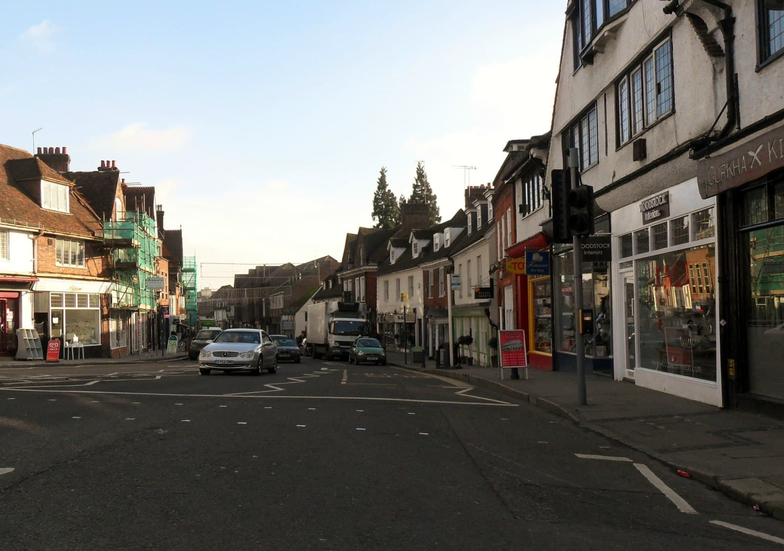 Sunlit high street lined with period shopfronts, with cars and a lorry on the road and scaffolding on the left