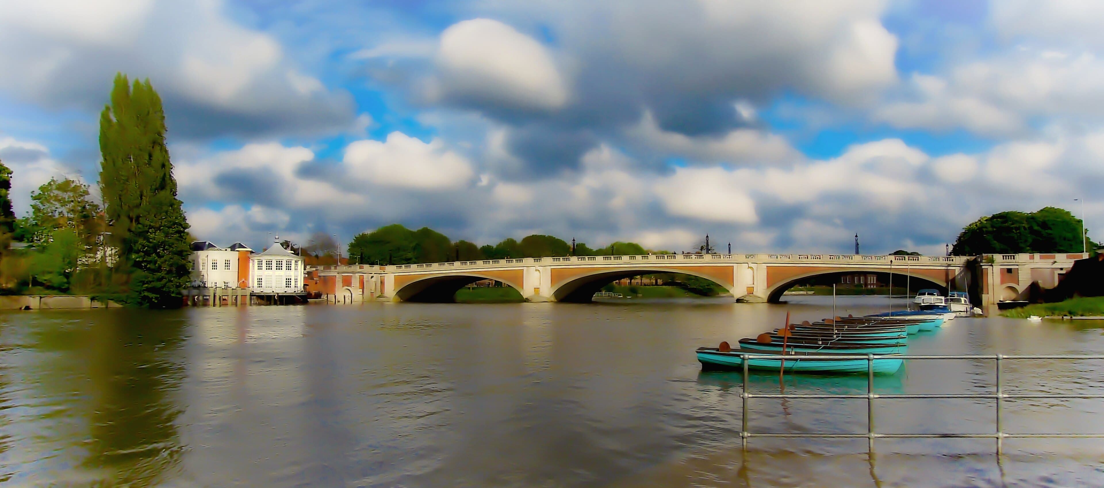 Hampton Court Bridge arching over the Thames beneath a dramatic cloudy sky, with turquoise rowing boats moored in the foreground