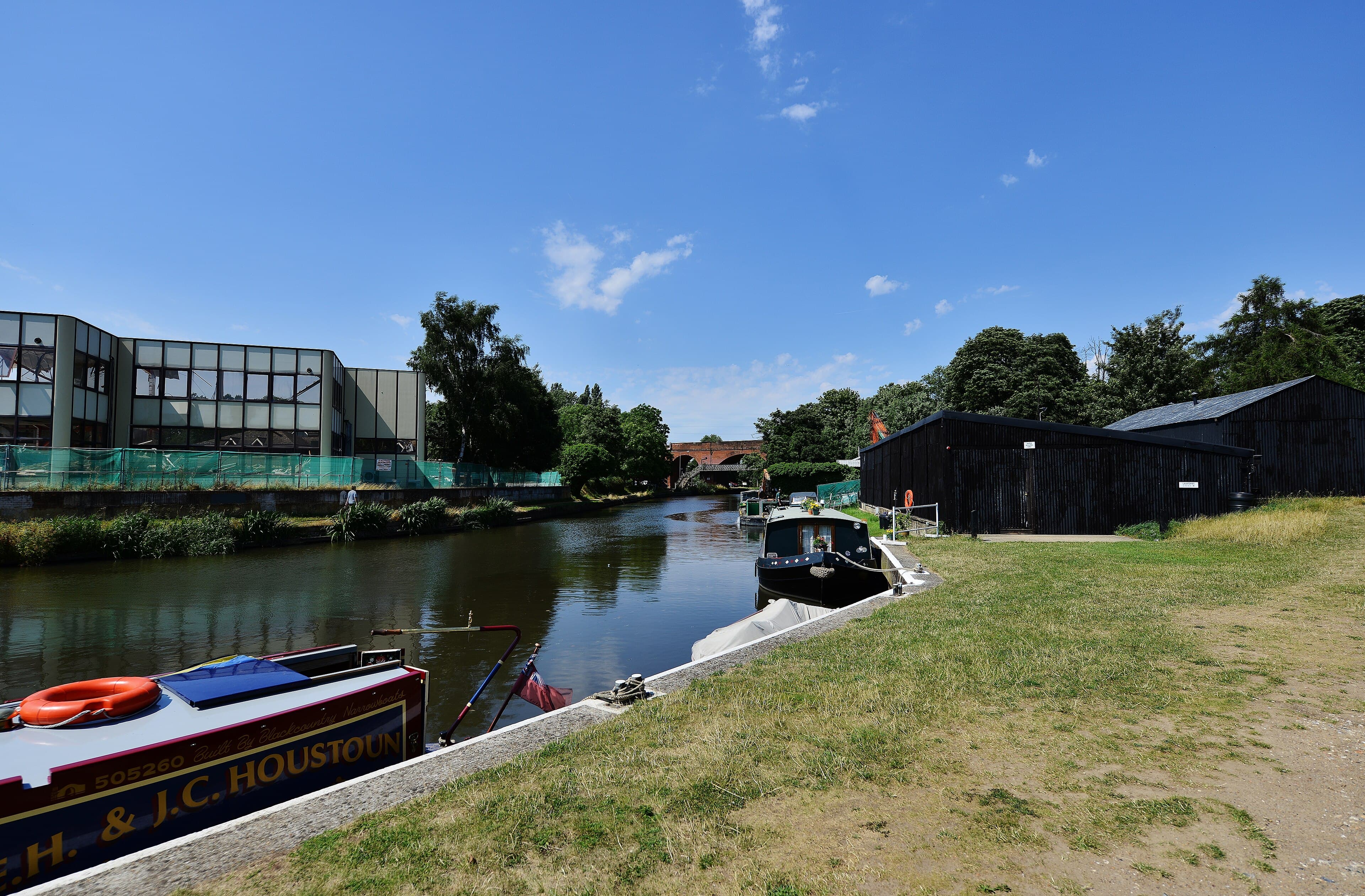 Narrowboats moored along a calm waterway beside a grassy bank, with a brick railway viaduct in the distance