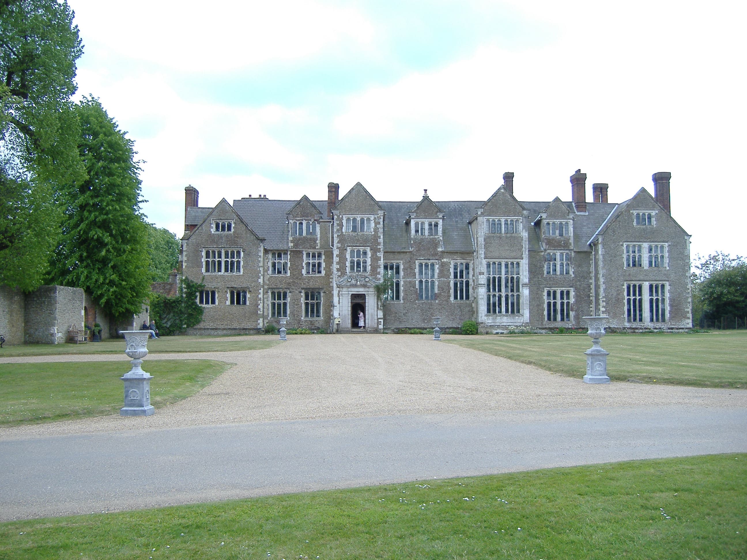Elizabethan stone manor house with gabled front, tall chimneys, gravel drive, stone urns and lawns under a pale sky