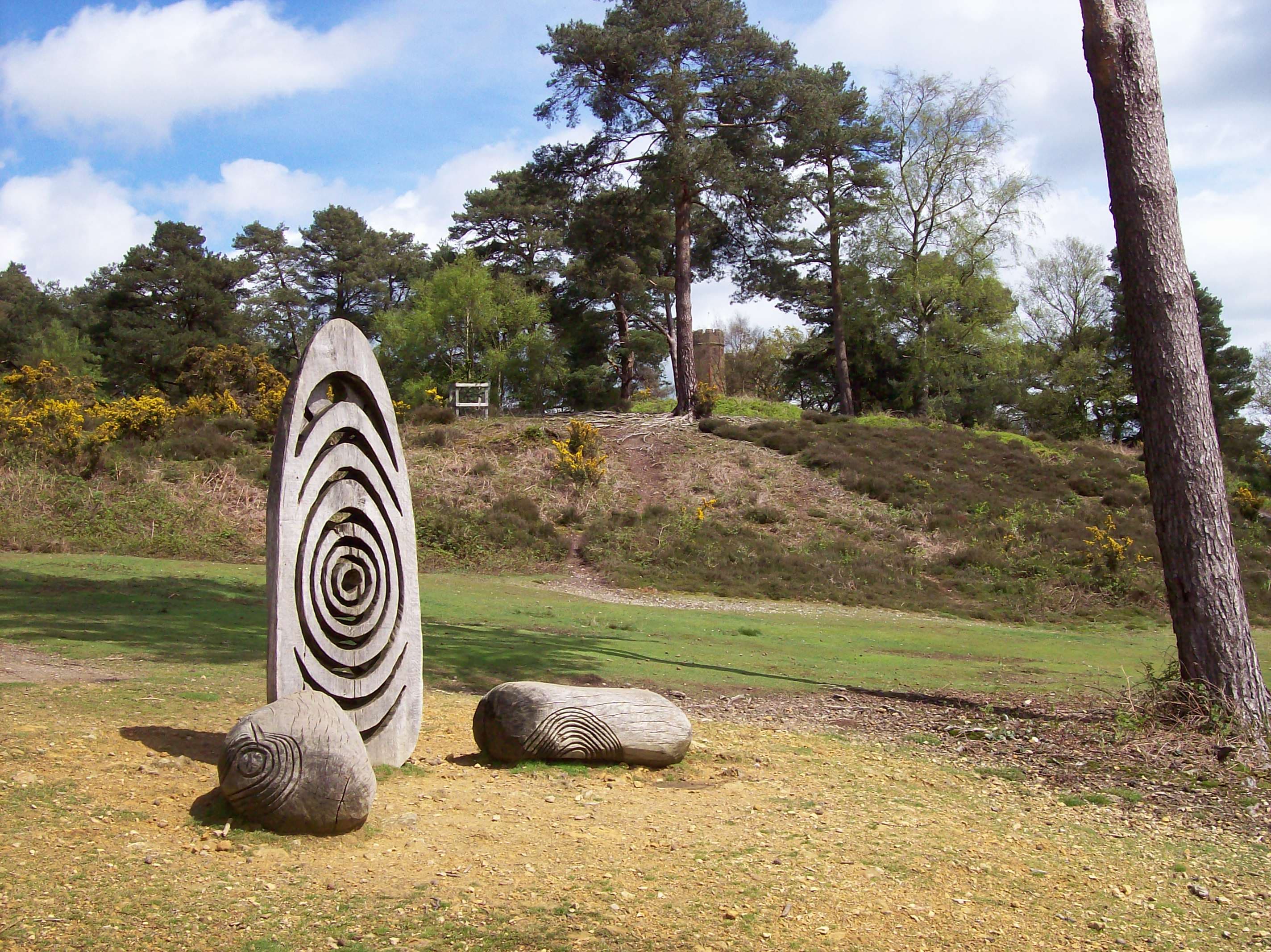 Carved wooden spiral sculptures on heathland below a pine-topped hill with a small stone tower beyond