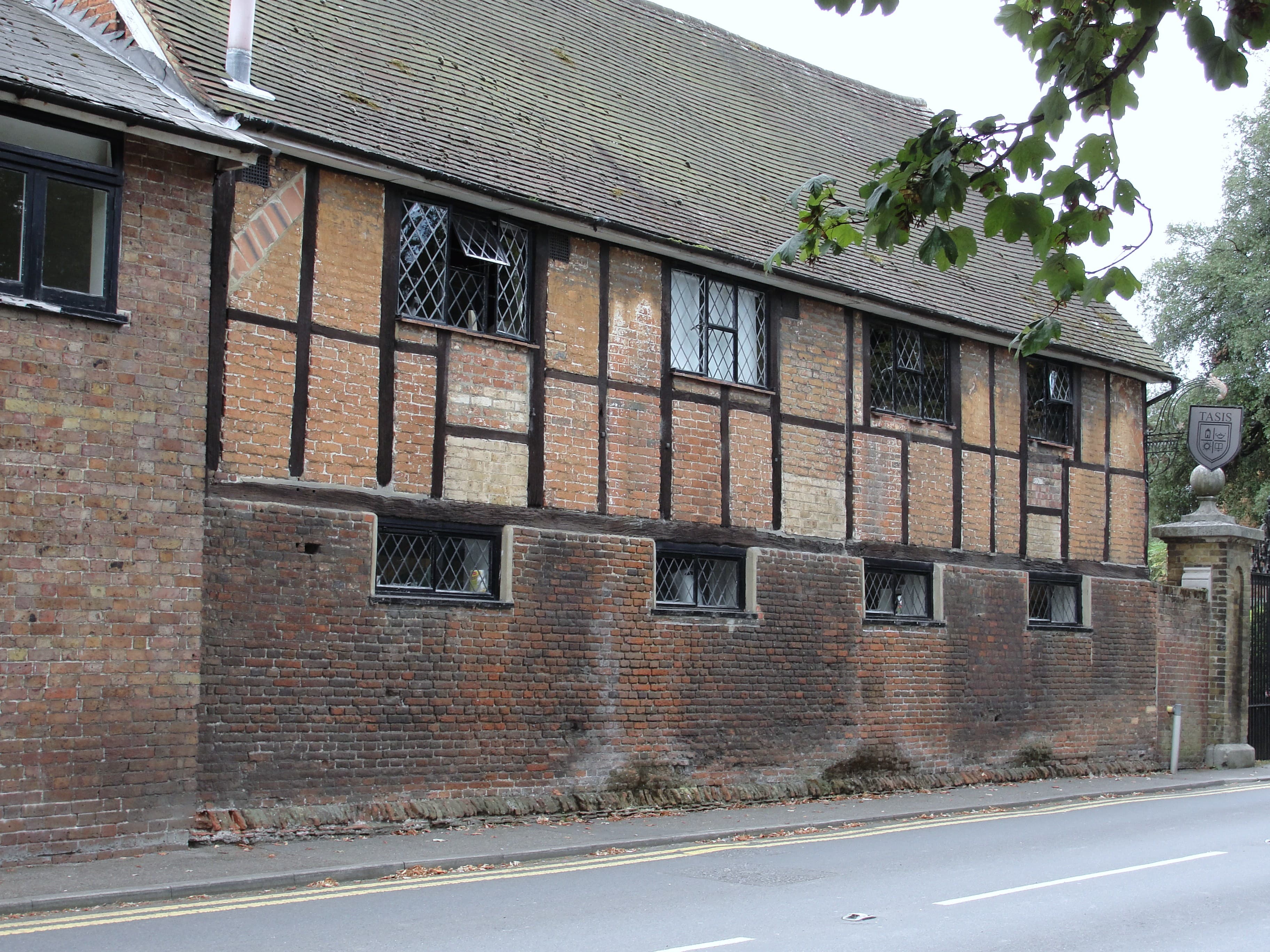 Old timber-framed building with brick infill and leaded windows beside a road, with a school gatepost nearby