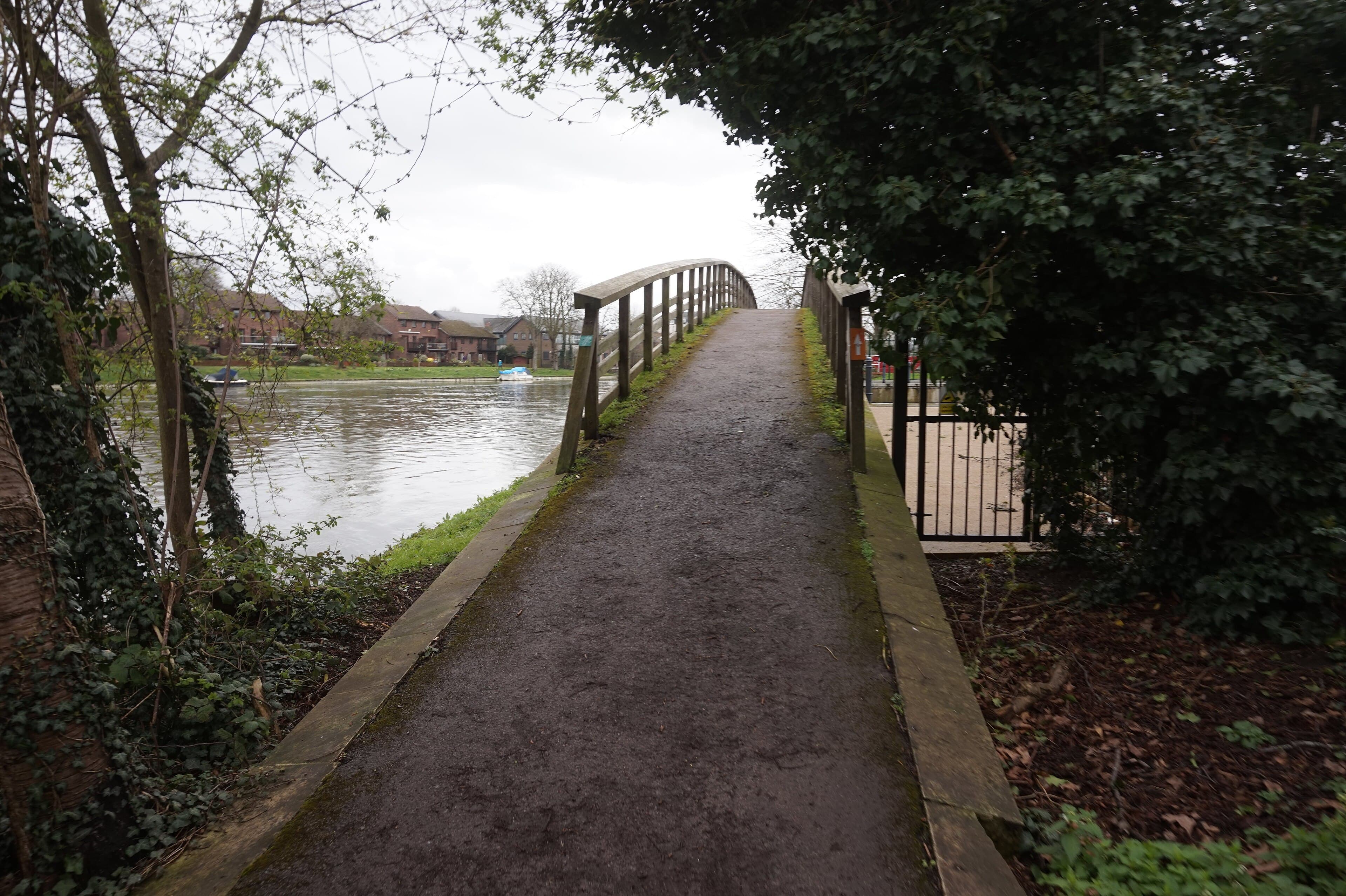 Arched wooden footbridge and tarmac towpath beside the River Thames, with riverside brick houses and moored boats opposite