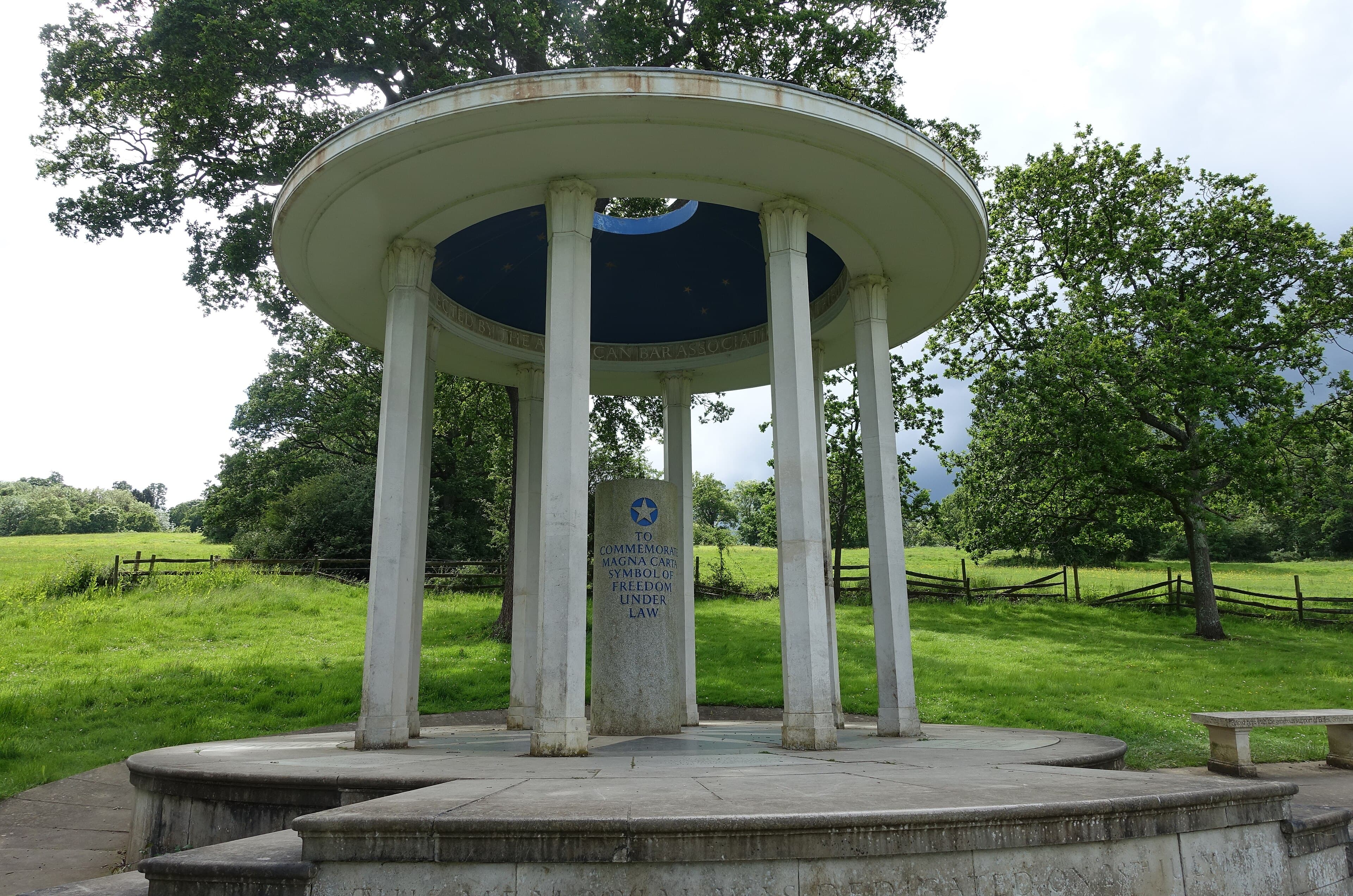 Circular white columned Magna Carta Memorial at Runnymede, with its inscribed stone pillar, surrounded by green meadow and oak trees