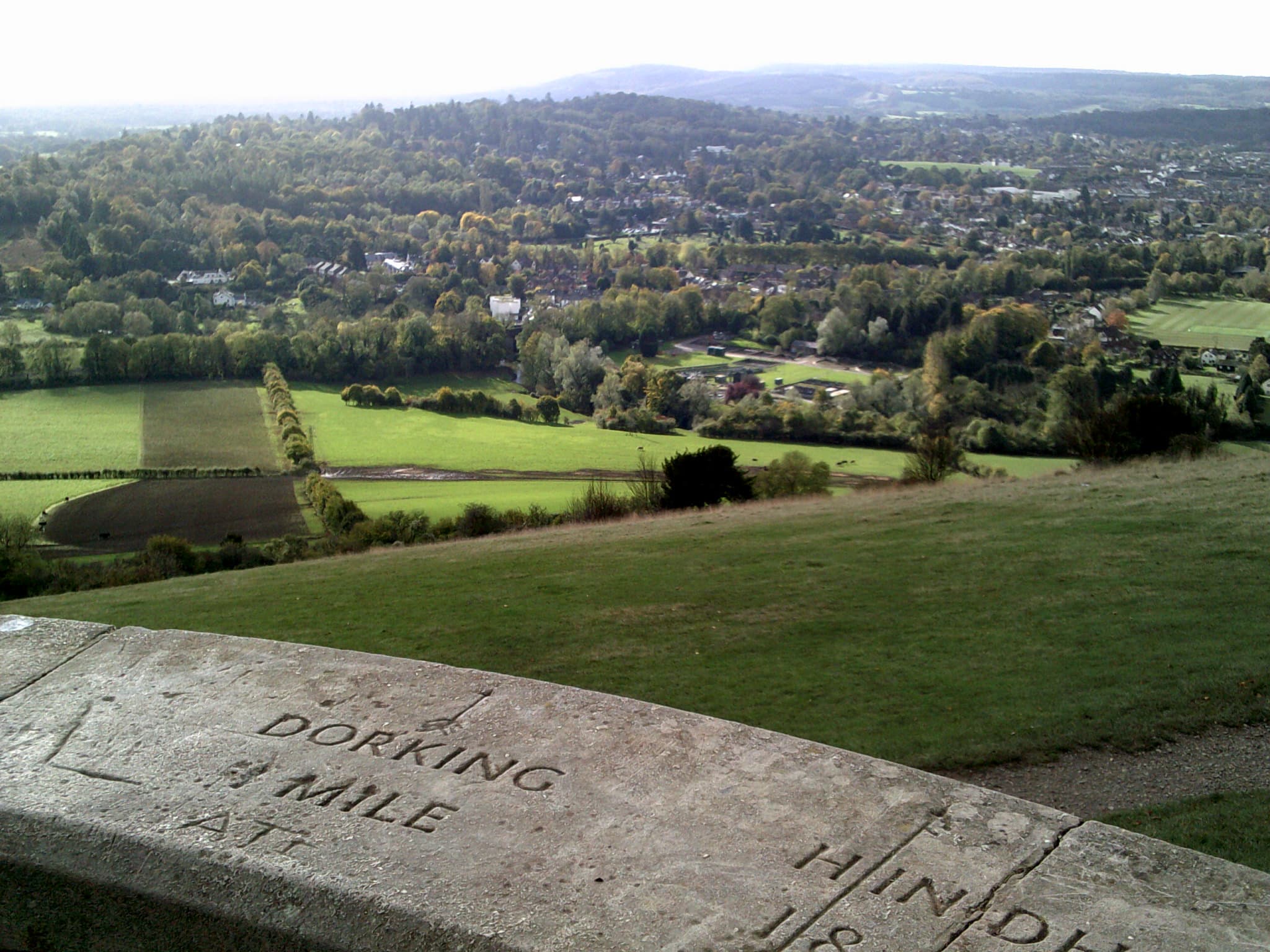 View over wooded Surrey countryside and fields from Box Hill viewpoint, with engraved stone indicator showing Dorking and Hindhead