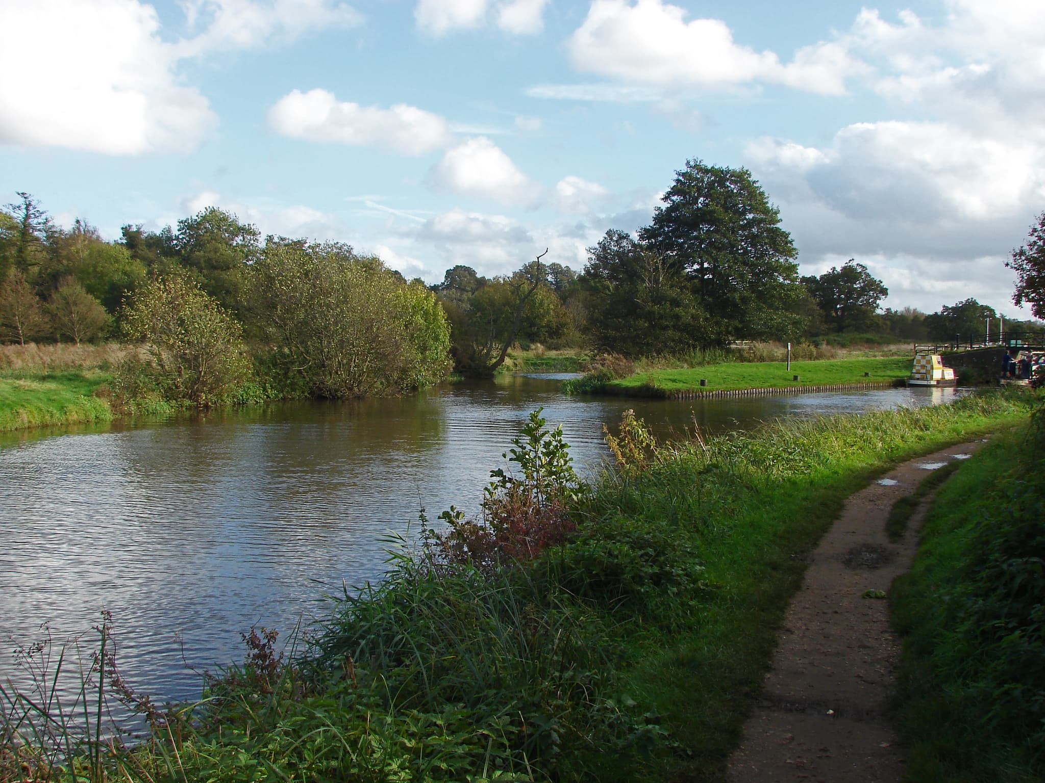 Towpath running beside a wide river with a narrowboat moored near a bridge under a partly cloudy sky
