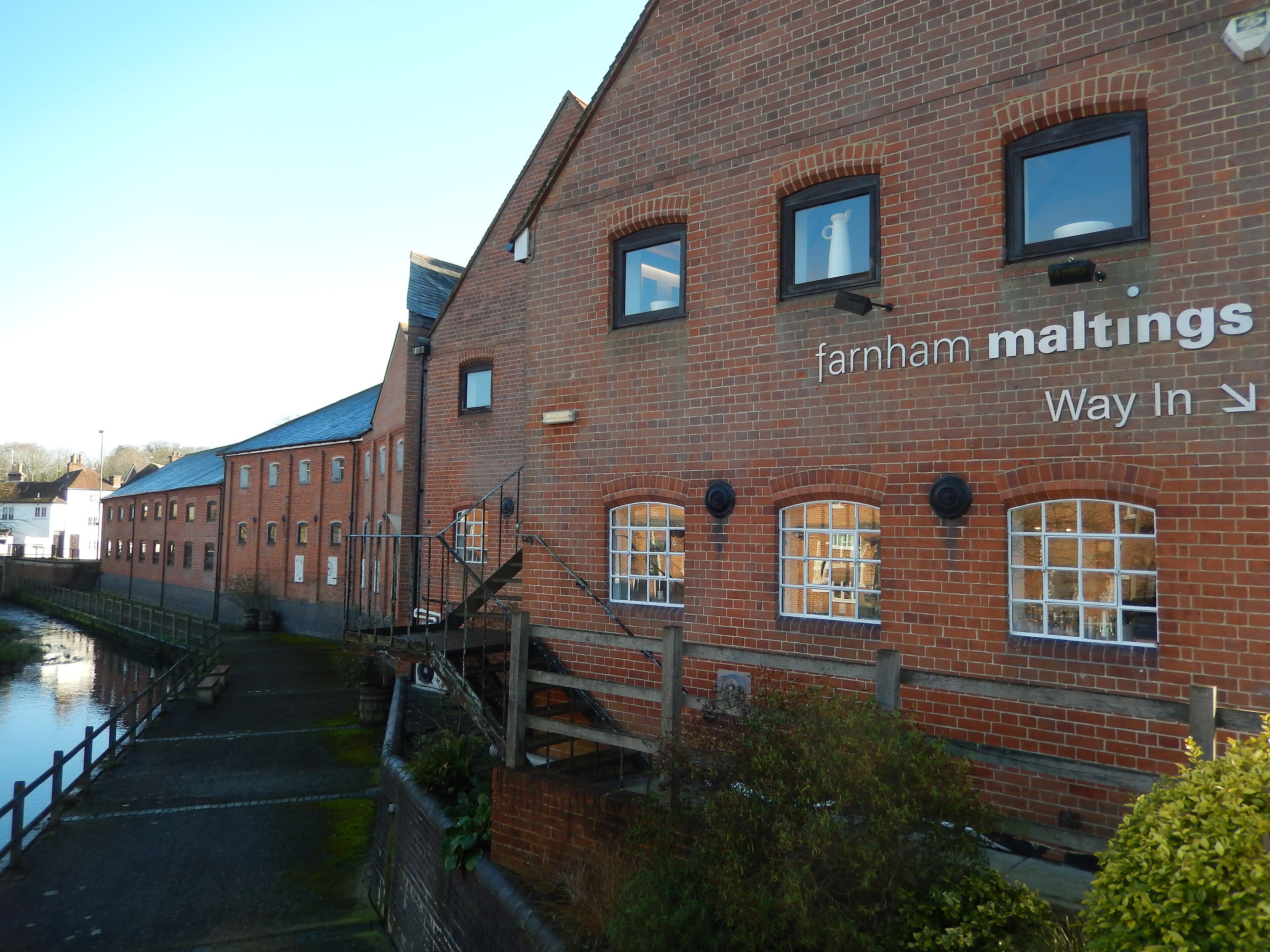 Red-brick Farnham Maltings building beside the River Wey on a clear day, with signage and a riverside path