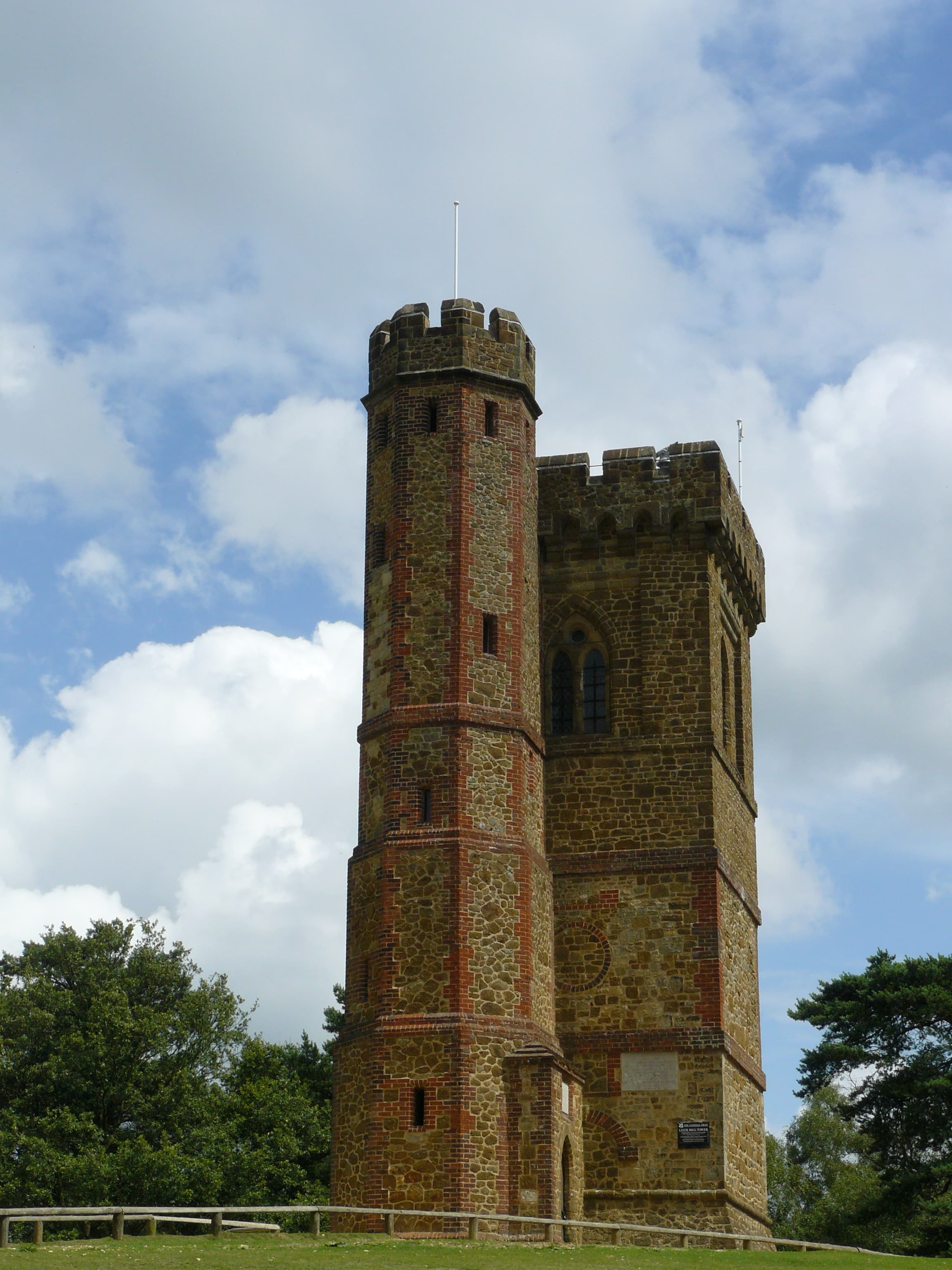 Crenellated stone and brick Gothic tower standing on a grassy hilltop beneath a bright blue sky with white clouds