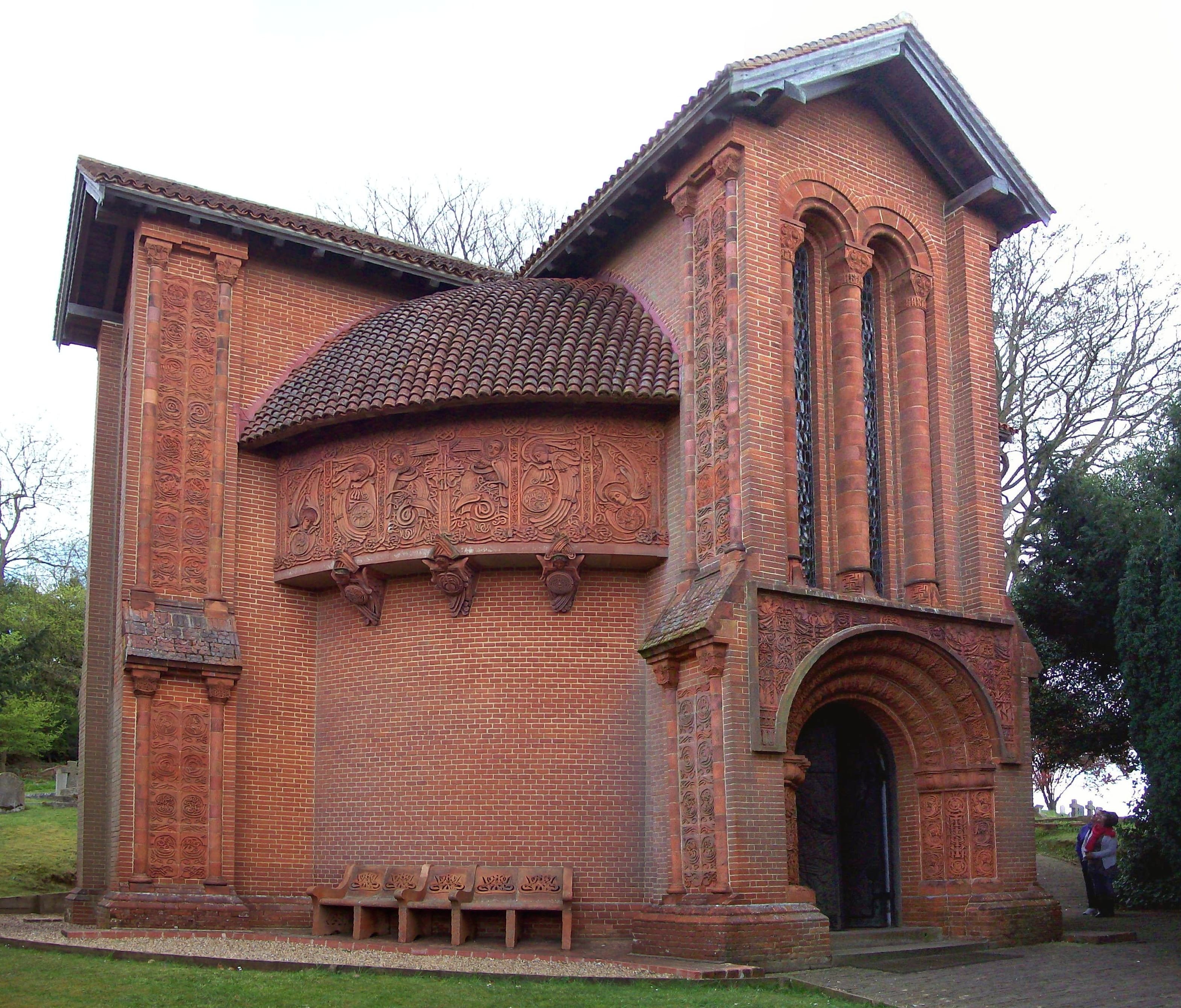Red brick and terracotta Watts Cemetery Chapel with ornate carved reliefs, arched doorway and tiled apse in a churchyard