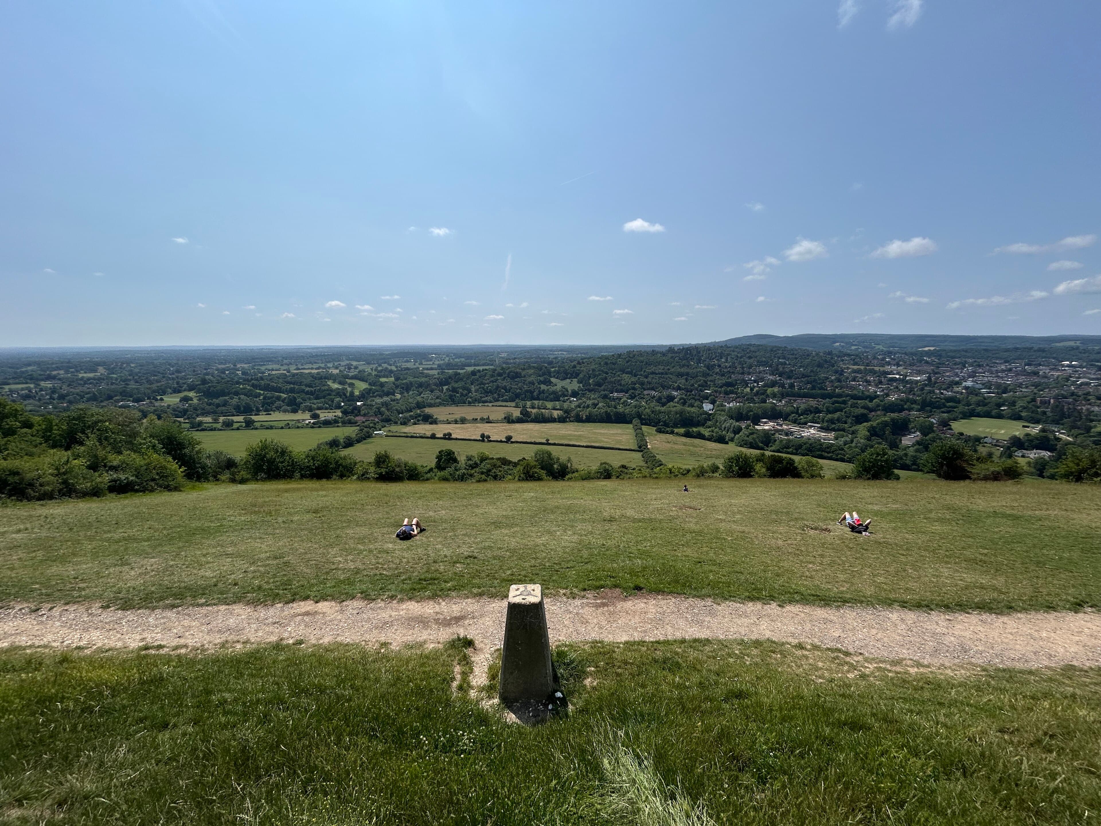 Trig pillar on a grassy hilltop with walkers resting and a wide view over wooded Surrey countryside