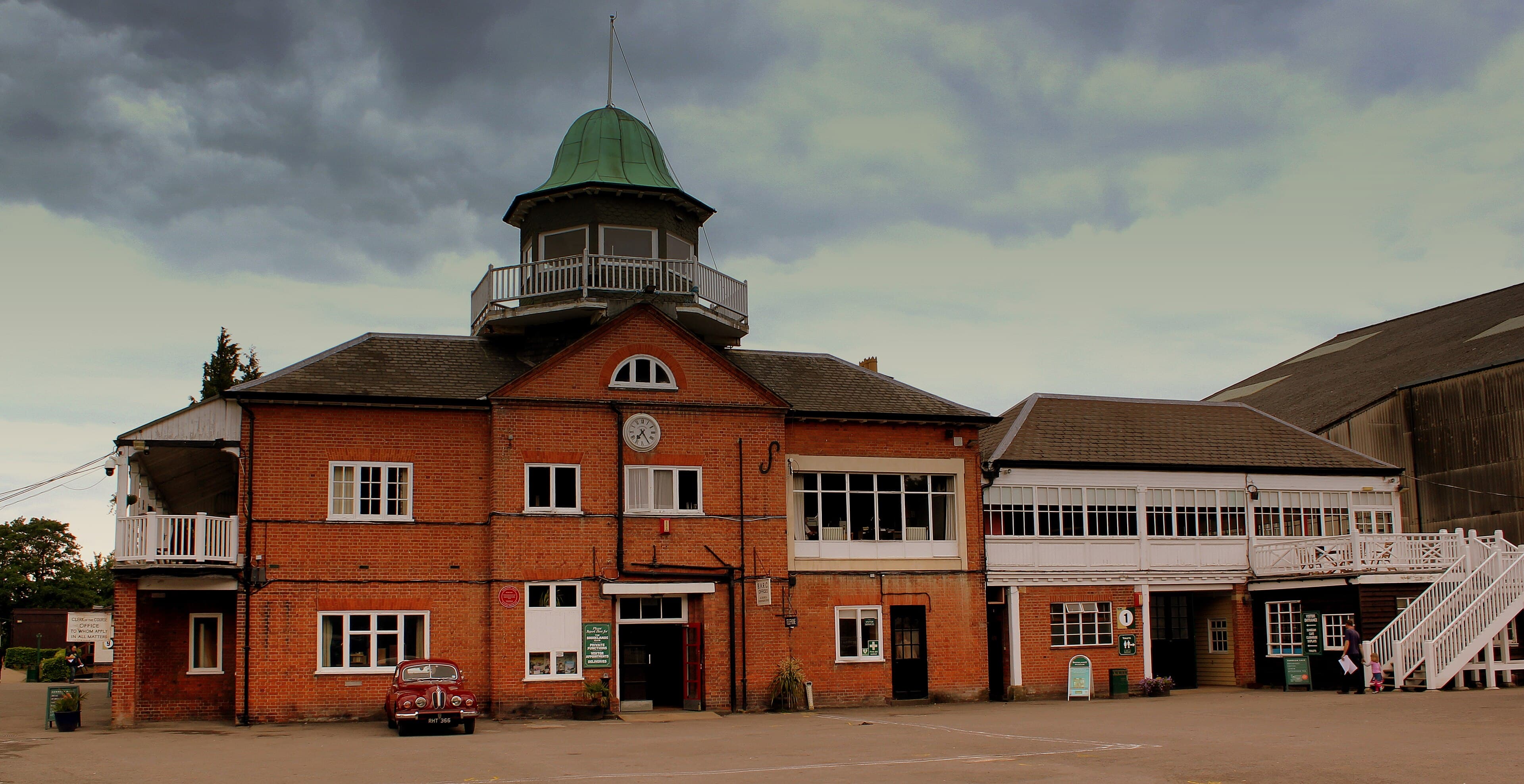 Brooklands clubhouse at Weybridge, red-brick with green-domed timing tower and vintage car parked outside